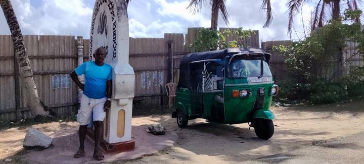 Man leaning on a roadside monument beside a green tuk-tuk under partly cloudy tropical skies.