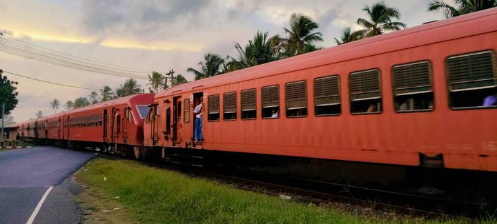 Bright red Sri Lankan train with a passenger hanging by the door, set against palm silhouettes at dusk.