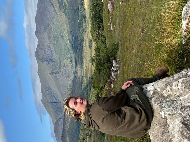 Traveller sitting on a rock overlooking expansive green valley and rugged Irish mountains.