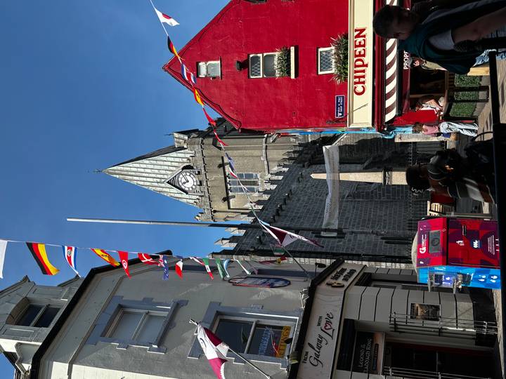 Colorful town street with medieval church tower and festive flags under bright blue sky.