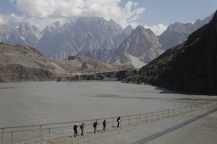 Small group crosses a narrow suspension bridge over a glacial lake amid jagged Karakoram peaks.