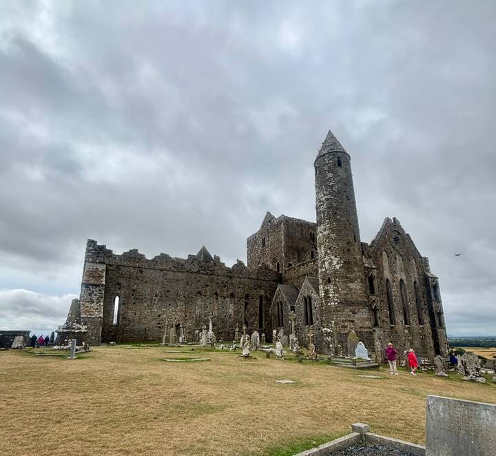 Medieval stone cathedral ruins on a hilltop with graveyard below under grey skies.