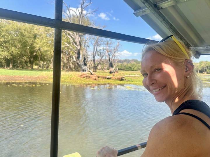 Woman smiling on a small boat cruising a calm lake with trees on shore.
