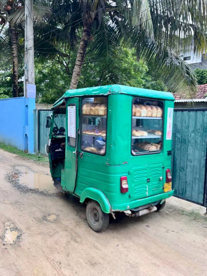 Green tuk-tuk bread van with shelves of loaves driving down a village lane.