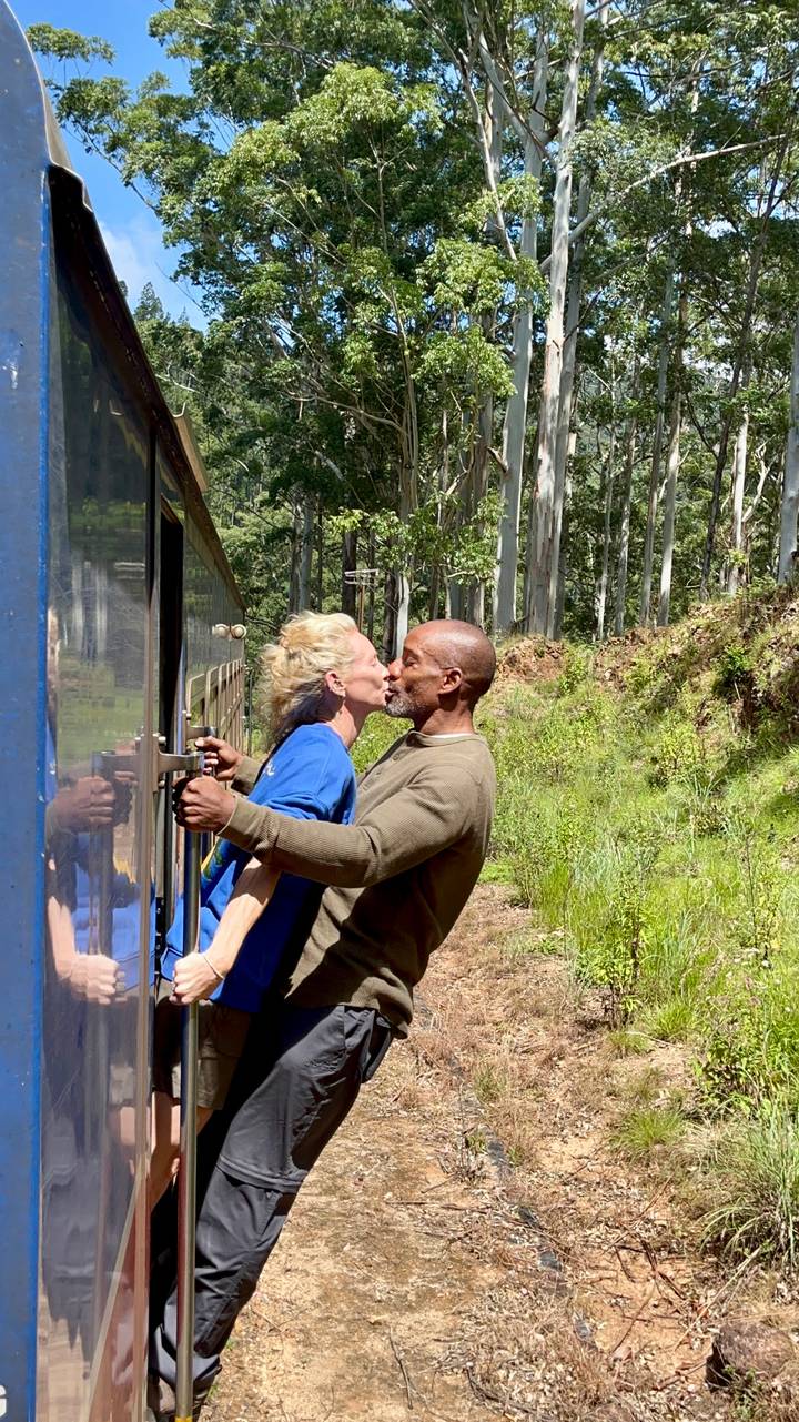 Man and woman lean out of the doorway of a blue train and share a kiss as the train moves through lush forest scenery.