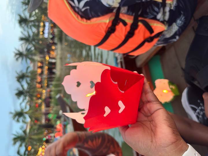 Hand holding a red paper lantern with heart cutouts on a wooden boat during Hoi An river lantern festival.