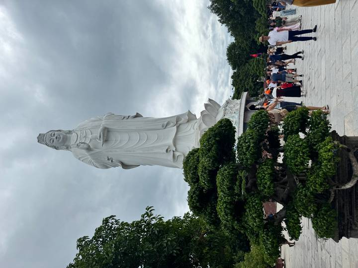 Tall white Lady Buddha statue towering over visitors in an open courtyard under cloudy skies.