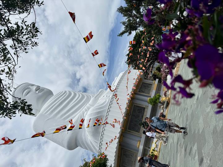 Giant seated Buddha statue with festive prayer flags and visitors walking below.