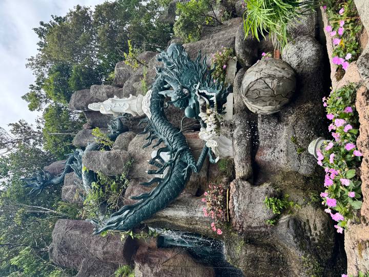 Intricate dragon and deity sculptures in rocky garden fountain with blooming flowers.