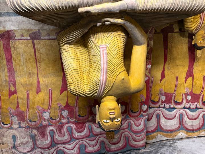 Illuminated golden Buddha statue seated before colorful mural figures inside a cave temple.