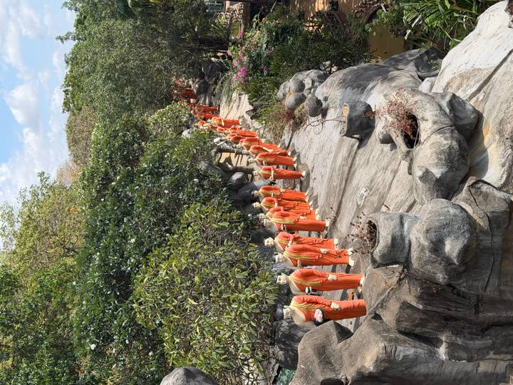 Long procession of orange-robed monk statues lines a rocky garden path beneath leafy trees.