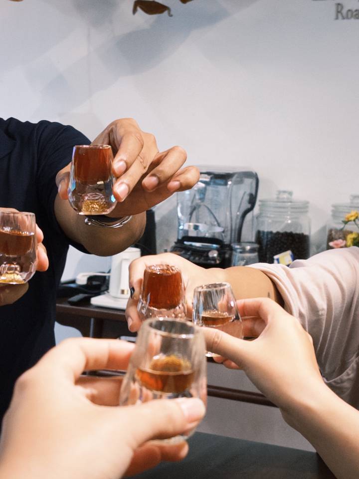 Group of hands toasting with small glasses of rich brown coffee liqueur in a cafe setting.