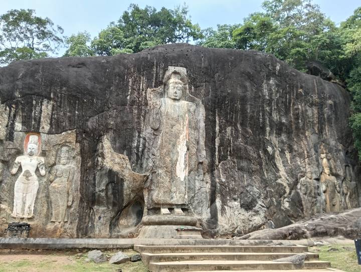 Ancient rock relief carvings depicting standing Buddhas etched into a massive dark cliff face in the jungle.