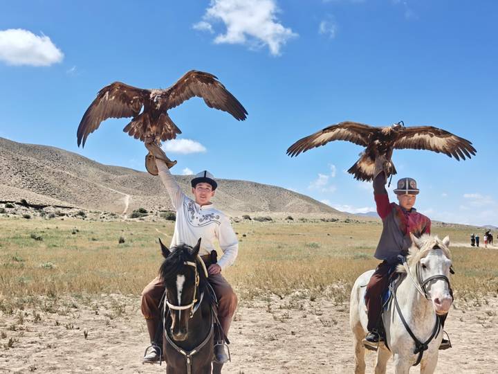Two horsemen in traditional attire lift golden eagles skyward on an open steppe under blue skies.