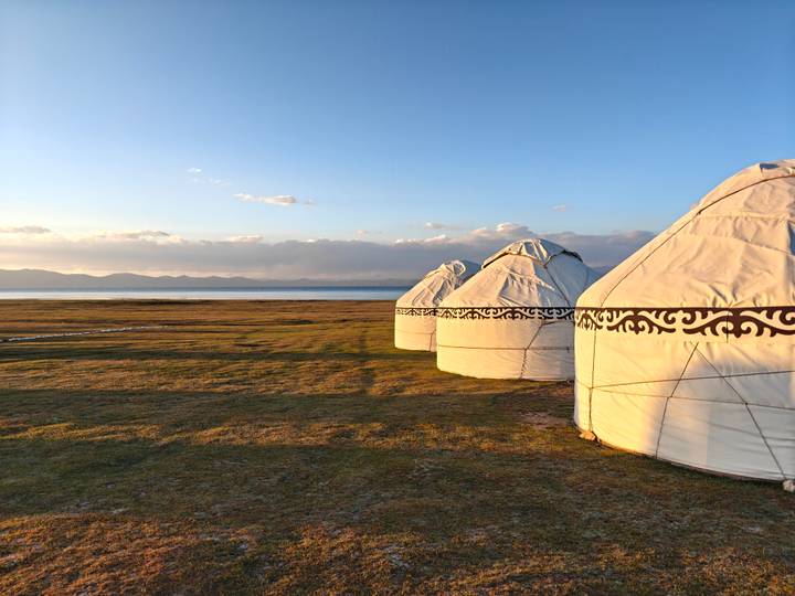 Row of white felt yurts beside a tranquil alpine lake during soft golden sunset light.