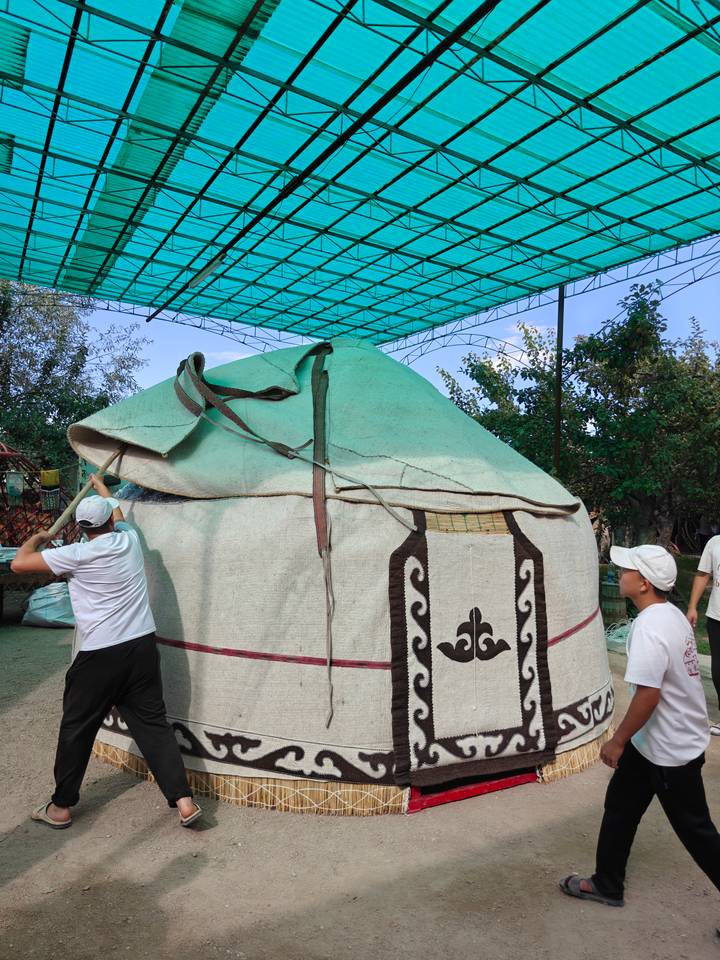 Two men assemble a traditional yurt frame under a shaded structure in a village yard.