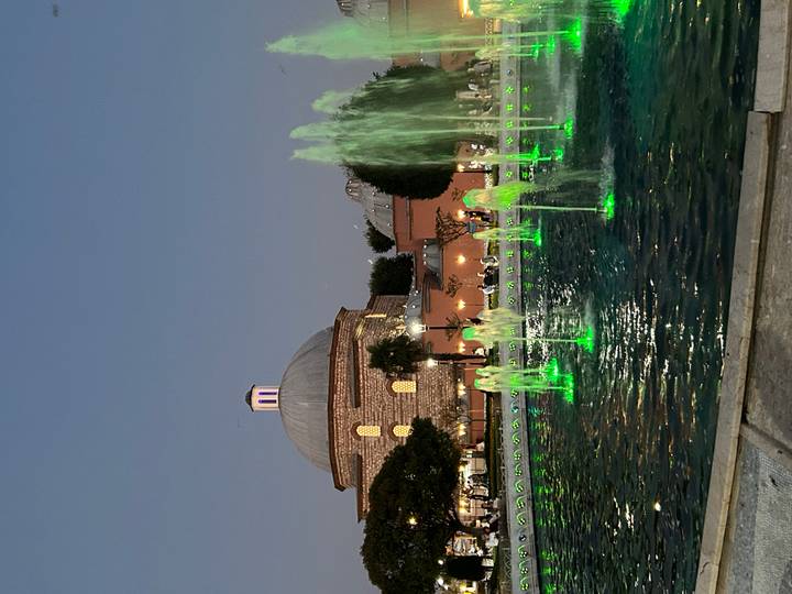 Evening scene of illuminated fountains glowing green before a domed historic bathhouse.