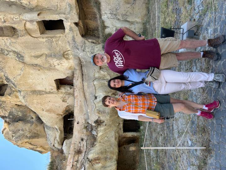Three smiling travellers pose before ancient cave dwellings carved into soft rock.