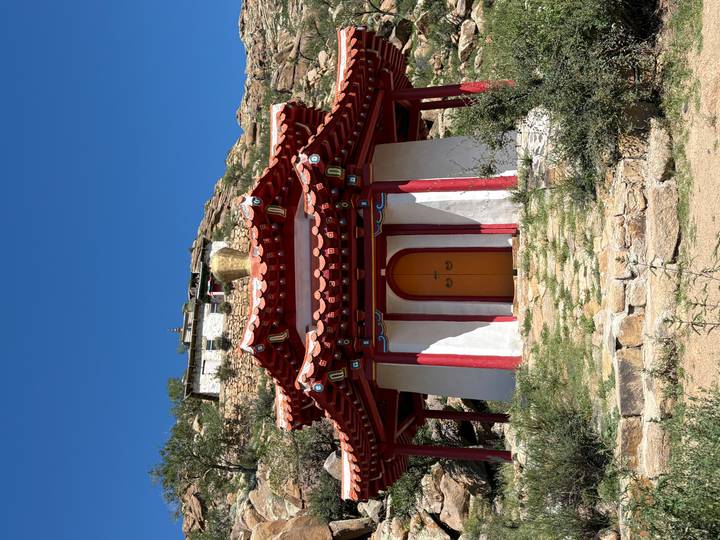 Small red-roofed temple with white walls and bright blue sky, perched on a rocky hillside.