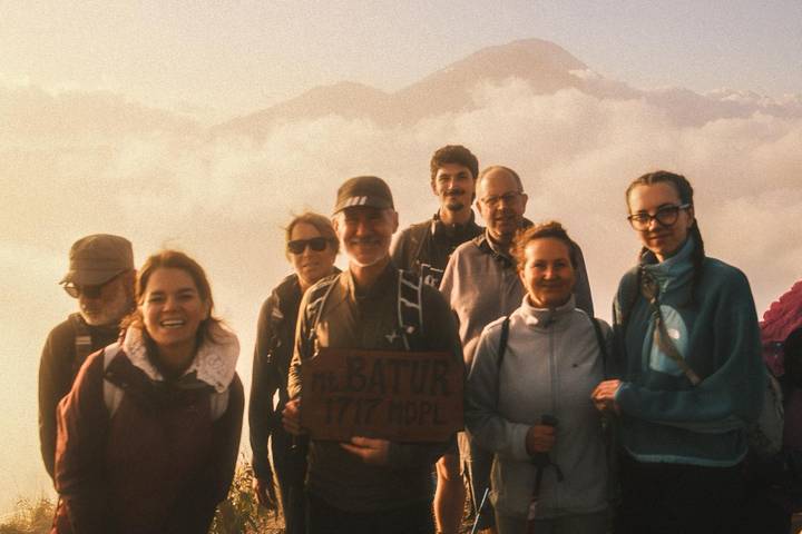 Hikers pose with a wooden Mt. Batur summit sign amid misty sunrise clouds.