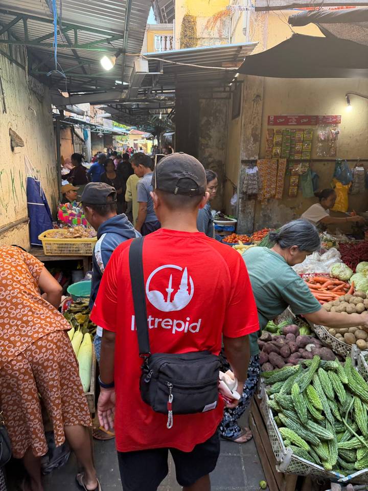 Bustling local market scene with an Intrepid guide in a red shirt leading travelers past fresh produce.