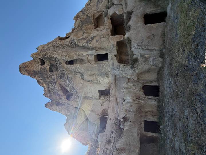 Sheer rock face riddled with ancient cave dwellings and dark openings under a clear blue sky in Cappadocia.