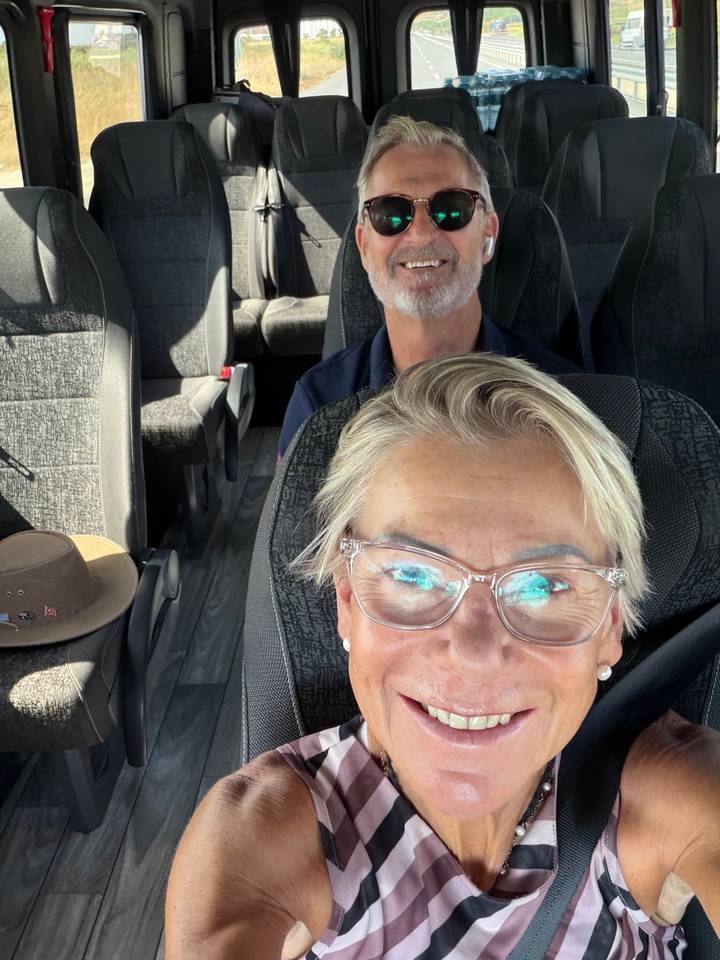 Close selfie of two travelers seated inside a tour van with rows of empty seats behind them.