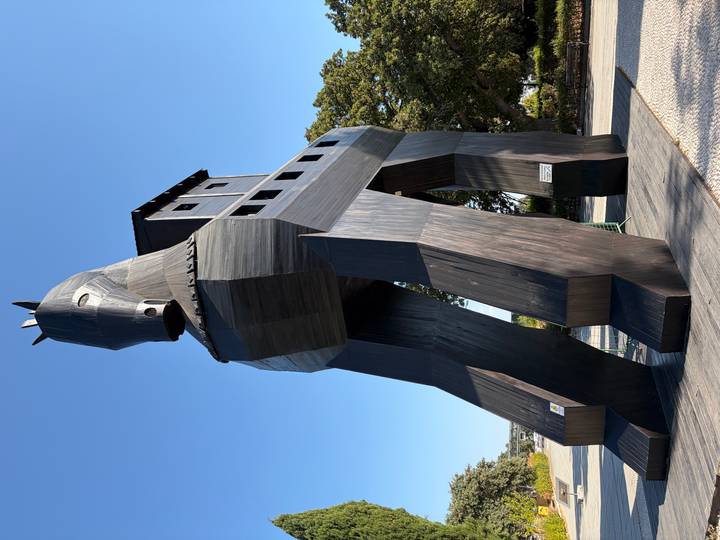 Large wooden replica of the Trojan Horse standing on a paved plaza against a clear blue sky.