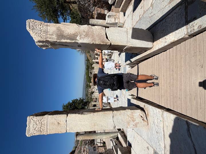 Visitor stretches arms between two tall marble pillars amid ancient stone ruins under a deep blue sky.