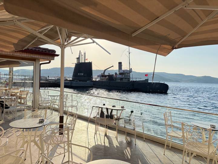 Black submarine moored beside an outdoor café on a sparkling waterfront with distant hills.