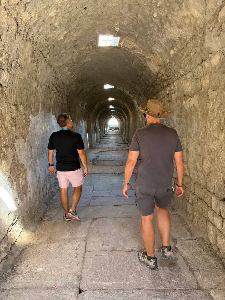 Two travellers walk through a long, stone-arched tunnel lit by daylight at the far end.