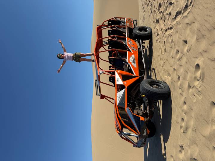 Traveler stands triumphantly on top of an orange dune buggy amid vast golden desert dunes.