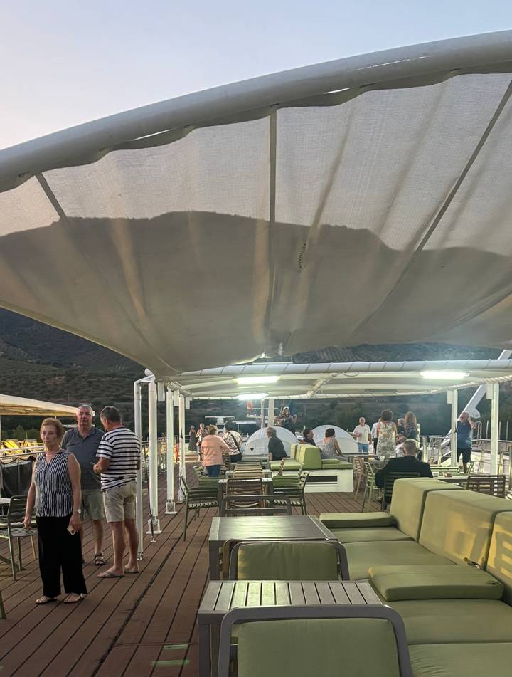 Evening gathering of cruise passengers relaxing on a covered sun deck with lounge seating.