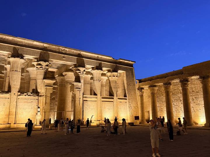 Nighttime scene of an illuminated columned Egyptian temple courtyard with tourists exploring.