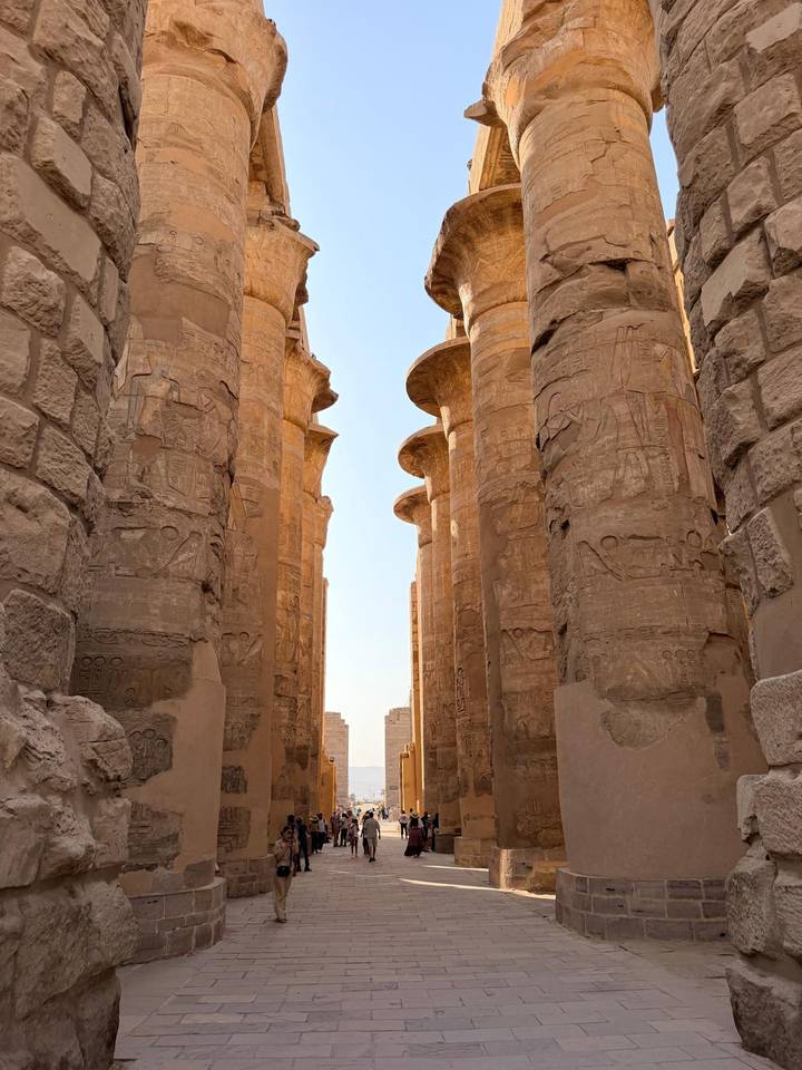 Massive sandstone columns adorned with carvings form a corridor in an ancient Egyptian temple.