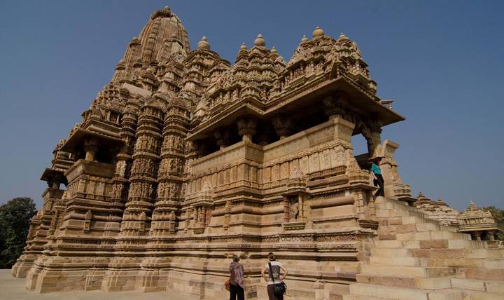 Visitors examining intricate sandstone carvings of a majestic Khajuraho temple under clear blue sky.