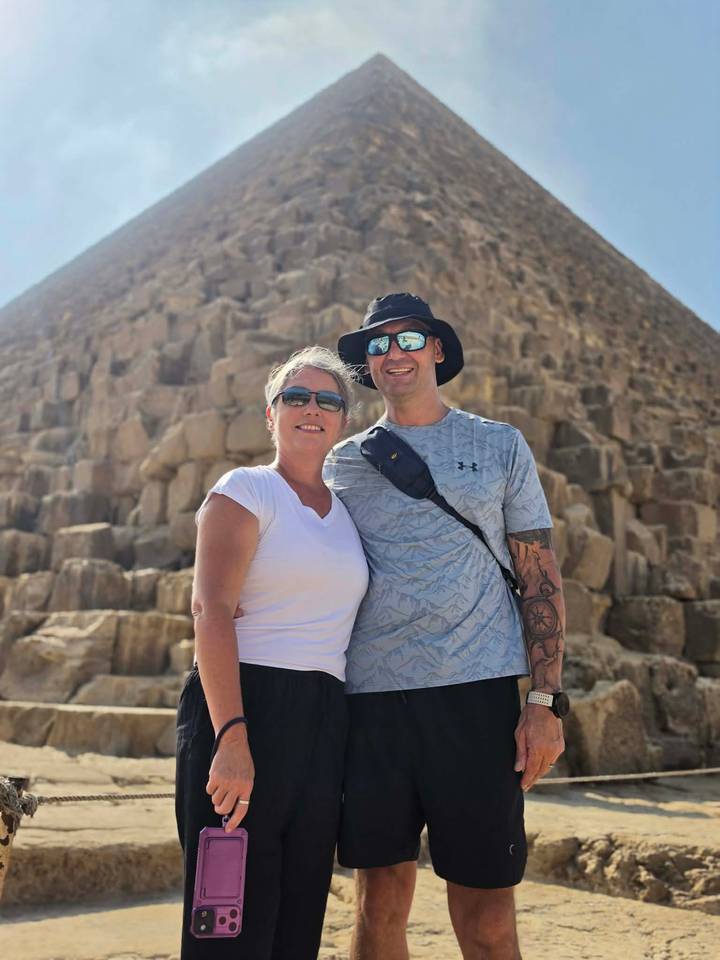 Couple pose in front of a pyramid’s limestone blocks wearing sunglasses and hats under bright sun.