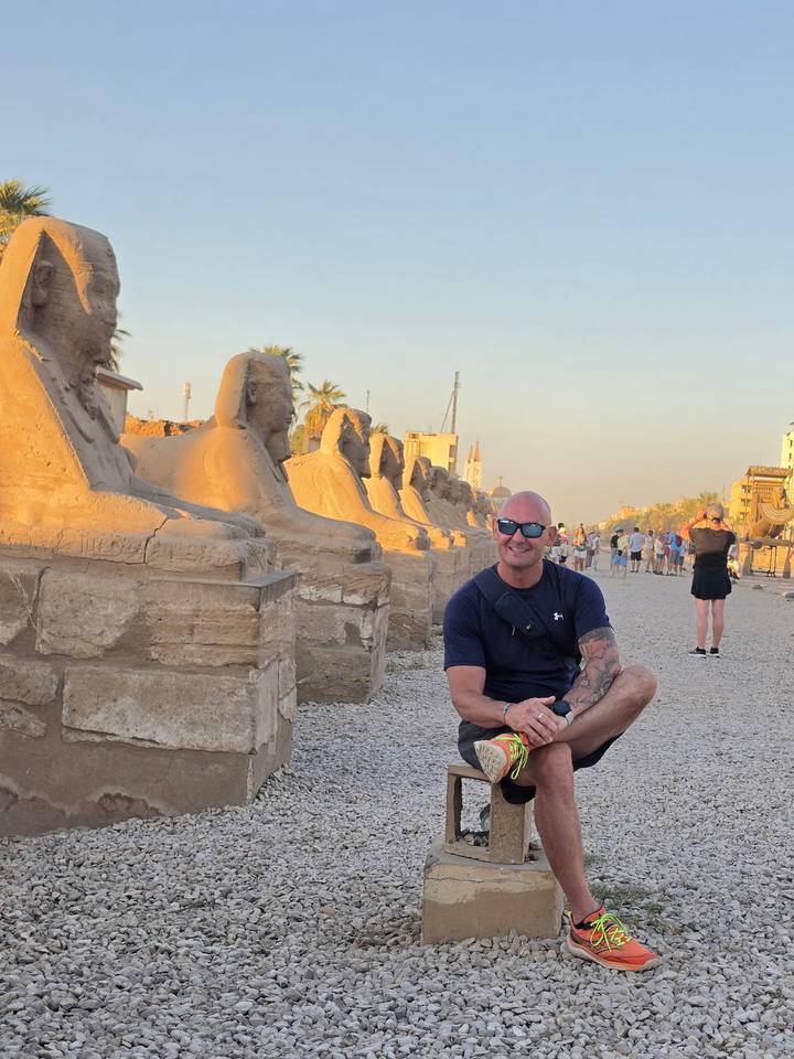 Man kneels for a photo beside a row of ram-headed sphinx statues on the Avenue of Sphinxes.