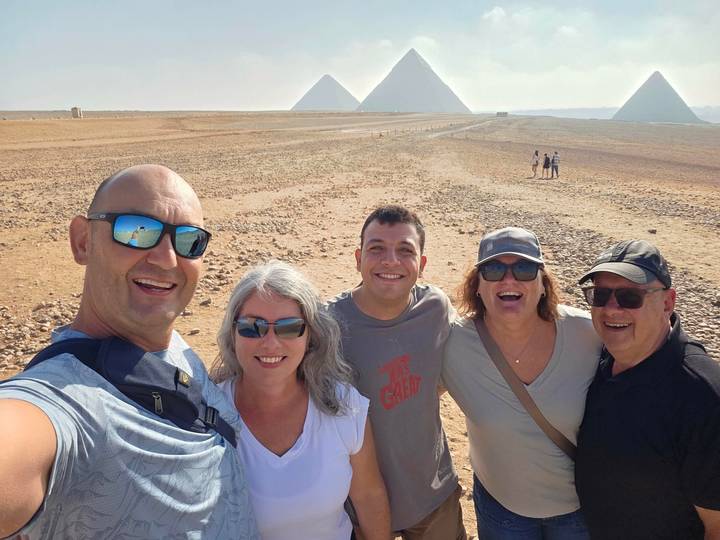 Group selfie in the desert with three pyramids visible on the distant horizon under bright sky.