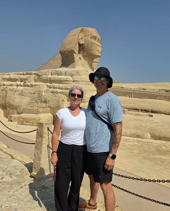 Smiling couple poses in front of the Great Sphinx and surrounding limestone ruins under a clear blue sky.