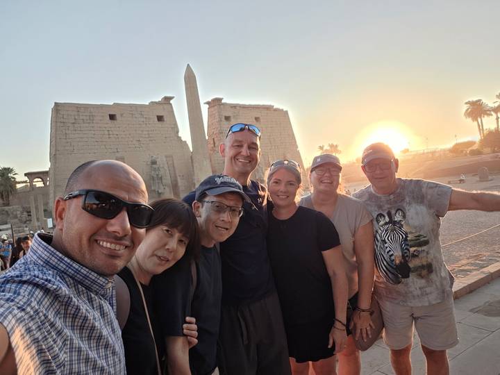 A diverse group of travelers takes a cheerful selfie at an ancient temple as the sun sets behind an obelisk.