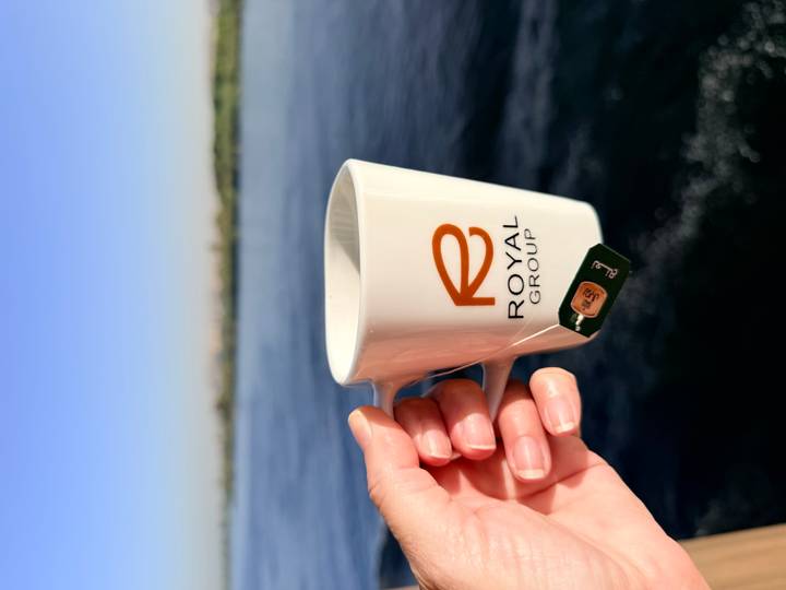Close-up of a hand holding a white Royal Group mug with water and distant riverbanks in soft morning light.
