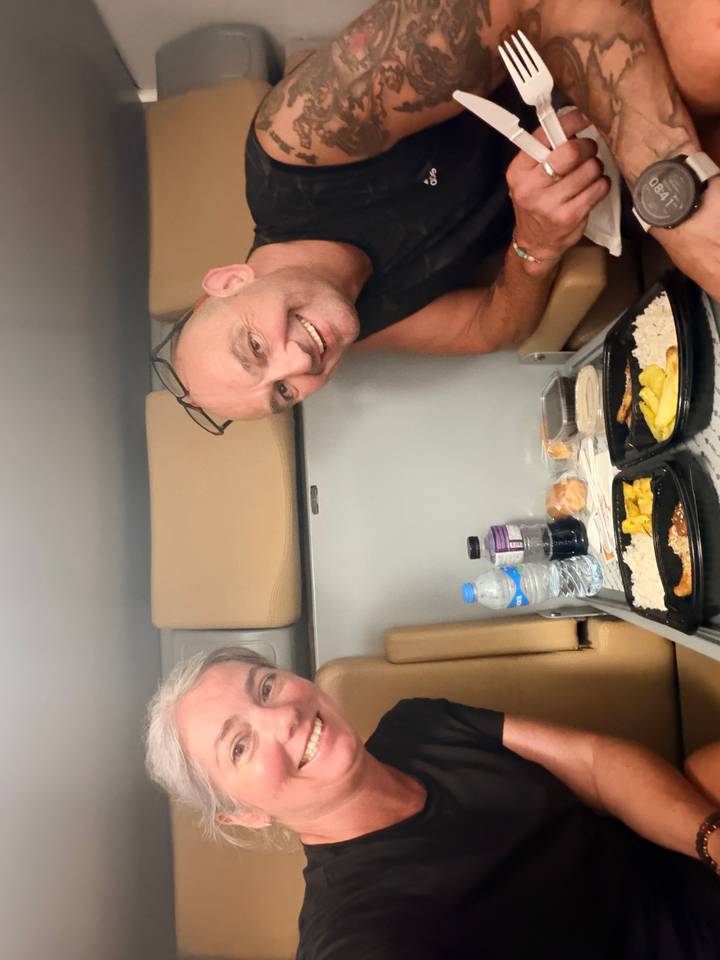 Two travelers enjoy a simple meal in a compact train cabin, smiling toward the camera.