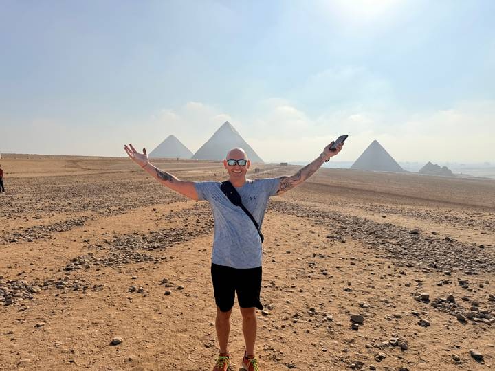 A traveler joyfully extends his arms on a vast desert plain with three pyramids in the distance.