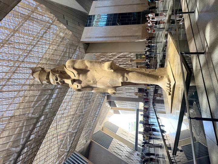 Colossal pharaoh statue stands beneath a dramatic glass ceiling inside a modern museum atrium.