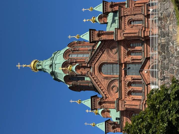 Brick-red Uspenski Cathedral with green copper domes and golden crosses rises against a cloudless blue Helsinki sky.