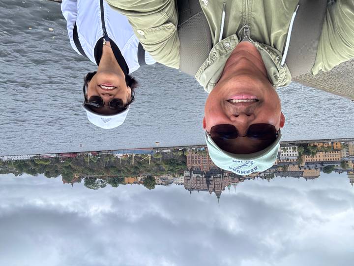 Couple smiles in selfie on Stockholm waterfront with historic hillside buildings under grey clouds.