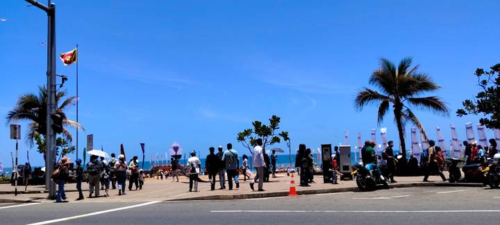 Busy seaside promenade with crowds, a Sri Lankan flag, palm trees and blue ocean beyond.