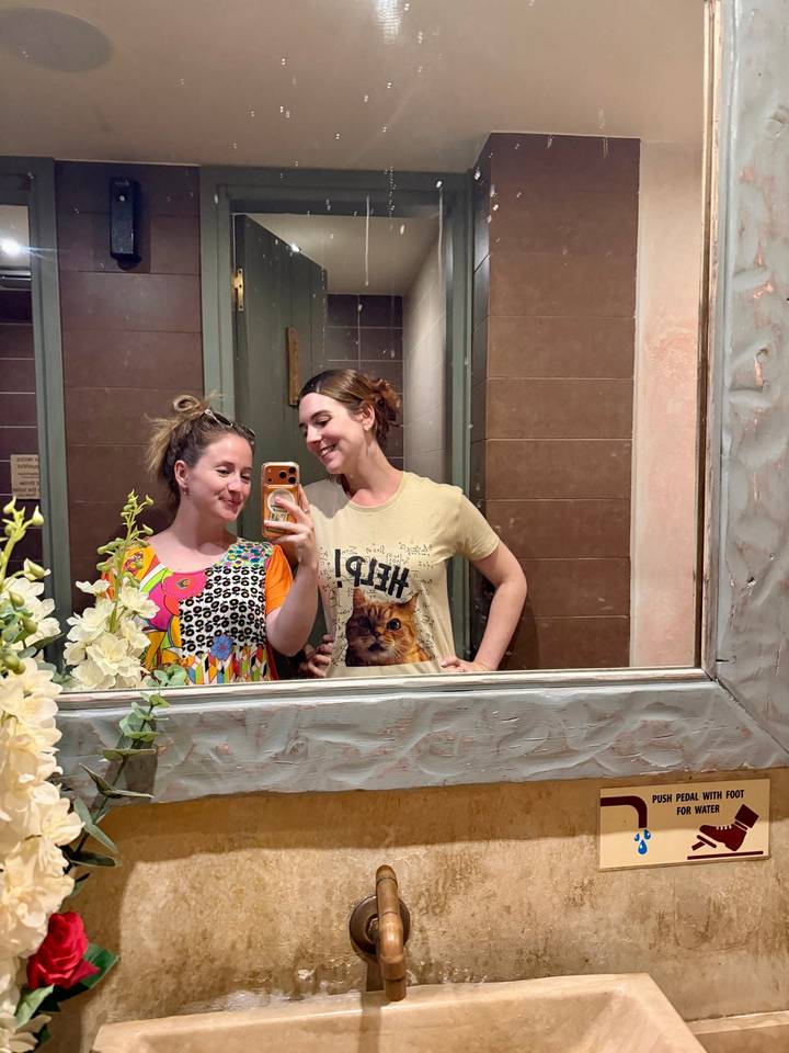 Two friends take a playful mirror selfie in a decorated restroom.