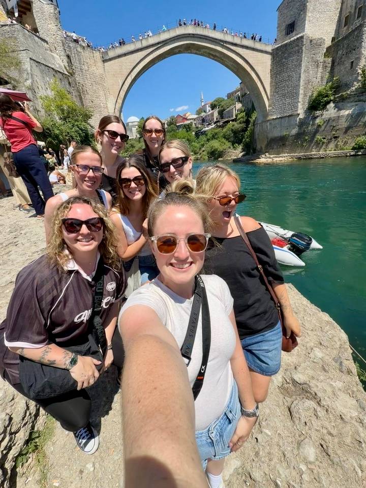 A cheerful group of young travellers taking a selfie on a stone riverside path beside clear turquoise water.